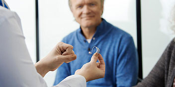 Billedet viser en hørespecialist, der holder et sæt høreapparater, så en mand og kvinde kan se dem.
