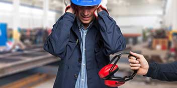 Billedet viser en mand på en arbejdsplads i støjende omgivelser. Han har en hjelm på og holder sig til ørerne.