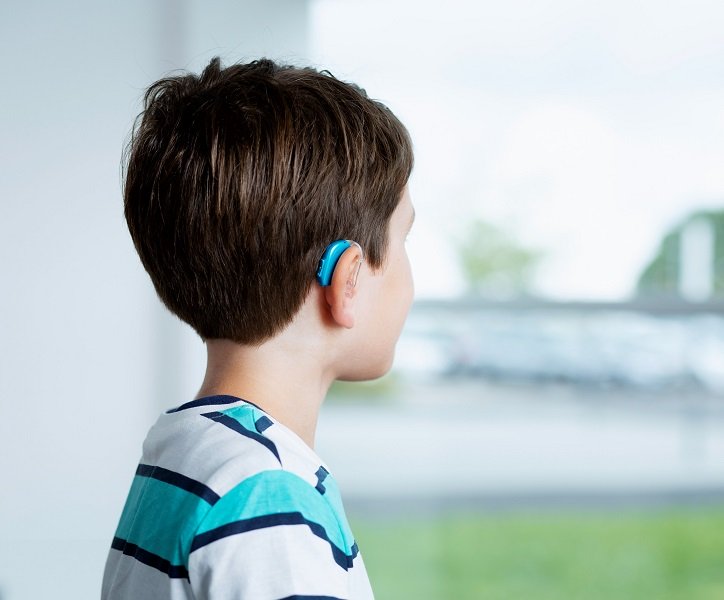 Dziecko z aparatem słuchowym (aparat słuchowy Oticon Opn Play) patrzy przez okno na zielony ogród.
