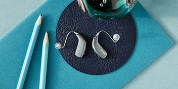 Hörapparater i färgen silver som ligger på ett mörkblått underlägg.