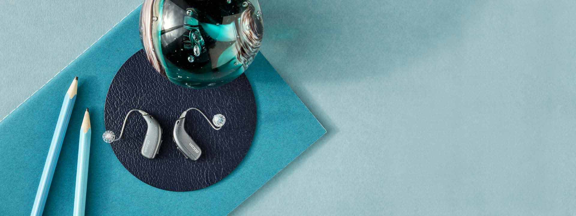 Två hörapparater på en mörkblå bakgrund. Bredvid hörapparaterna ligger två pennor.