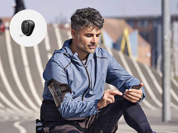 Bild på en man som sitter och tittar ner i sin telefon som har en Oticon Own hörapparat, bredvid kan man se hur en Oticon hörapparat ser ut
