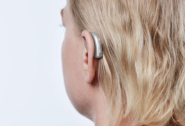 Hörapparat som finns att prova i Östergötland. Bärs bakom örat av en blond kvinna. Bilden på bakhuvudet och örat.