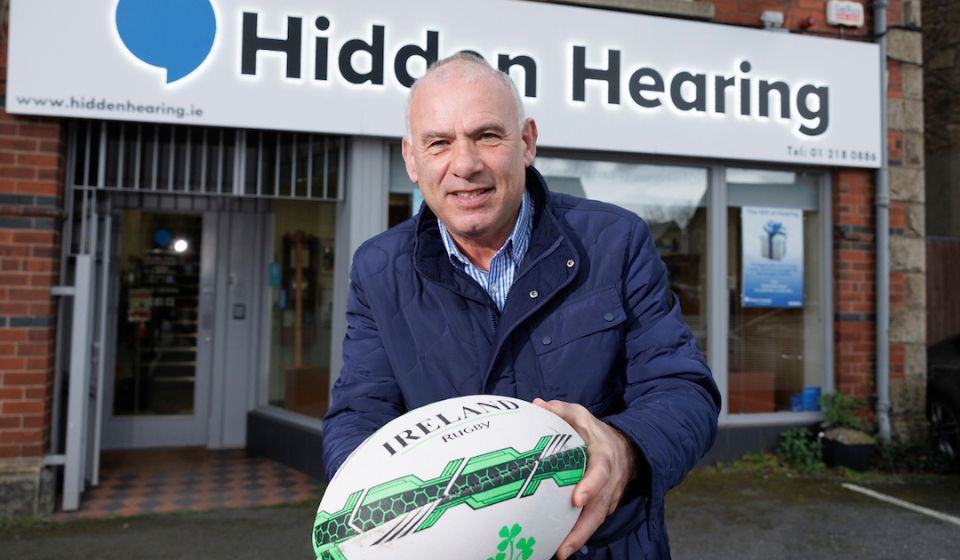 Tony Ward holding out a rugby ball while standing in front of a hidden hearing clinic