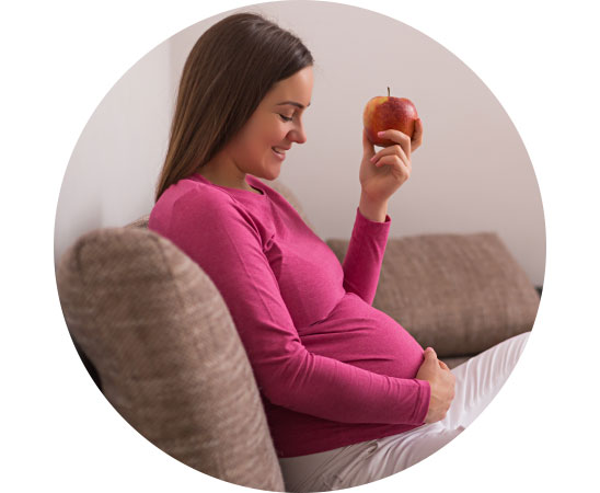 La imagen muestra una mujer embarazada sentada en el sofá comiendo una manzana