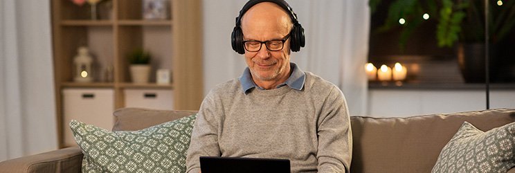 Image shows man sitting on sofa with laptop and headphone on